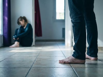 Bare feet in the foreground while a woman sits huddled against a wall down the hallway